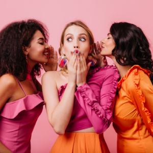 Tres mujeres en un fondo rosa, dos de ellas están susurrando a la mujer del centro
